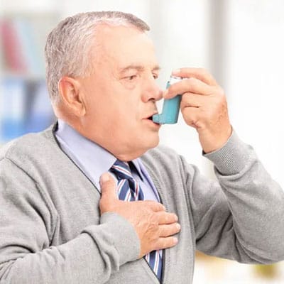 Elderly man using an inhaler, with one hand on his chest, in a well-lit room.