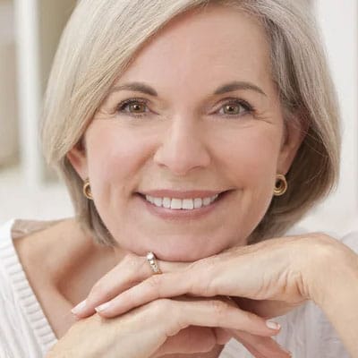 A smiling woman with short gray hair rests her chin on her hands. She is wearing a white top and gold hoop earrings.