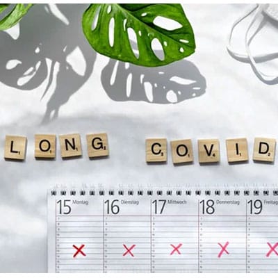 Wooden letter tiles spell "Long COVID" above a calendar with red X marks. A green plant and a face mask are nearby.