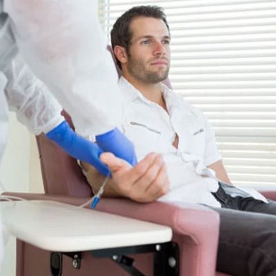 A person in a white shirt sits in a chair, receiving an injection from a healthcare worker wearing blue gloves.