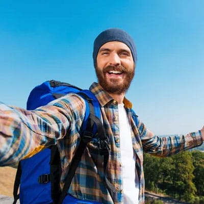 A bearded man wearing a beanie and plaid shirt takes a selfie outdoors with a blue backpack on. Clear blue sky and greenery are in the background.