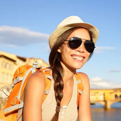 Woman in a hat and sunglasses smiles outdoors, wearing a beige top and an orange backpack. Bright, sunny day with a clear blue sky in the background.