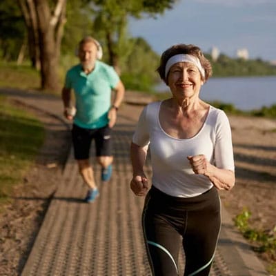 Two people jogging on a sunny path near a lake, with trees and distant buildings in the background. The woman in the foreground is wearing a white headband and top.
