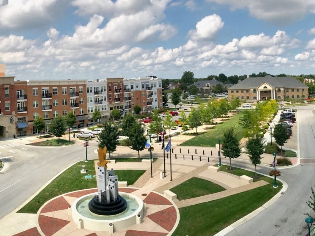 Aerial view of a town square with a modern sculpture, surrounded by brick buildings, a parking area, and greenery. Cloudy sky in the background.