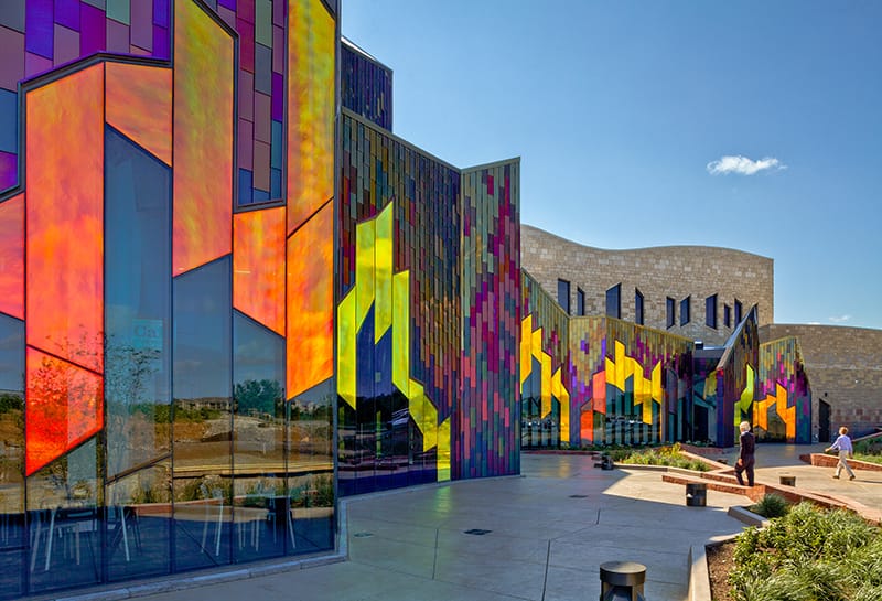 A modern building with multicolored, geometric stained glass panels reflecting sunlight against a clear blue sky. Two people are walking in front of the building.