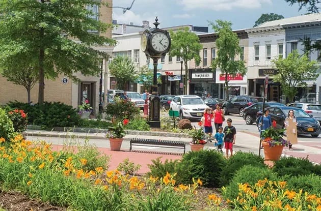 A town square with a large clock, people walking, benches, and flowerbeds. Shops and parked cars line the street under a clear sky.