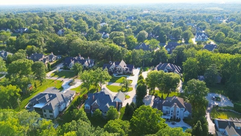 Aerial view of a suburban neighborhood with large houses surrounded by lush green trees under a clear blue sky.