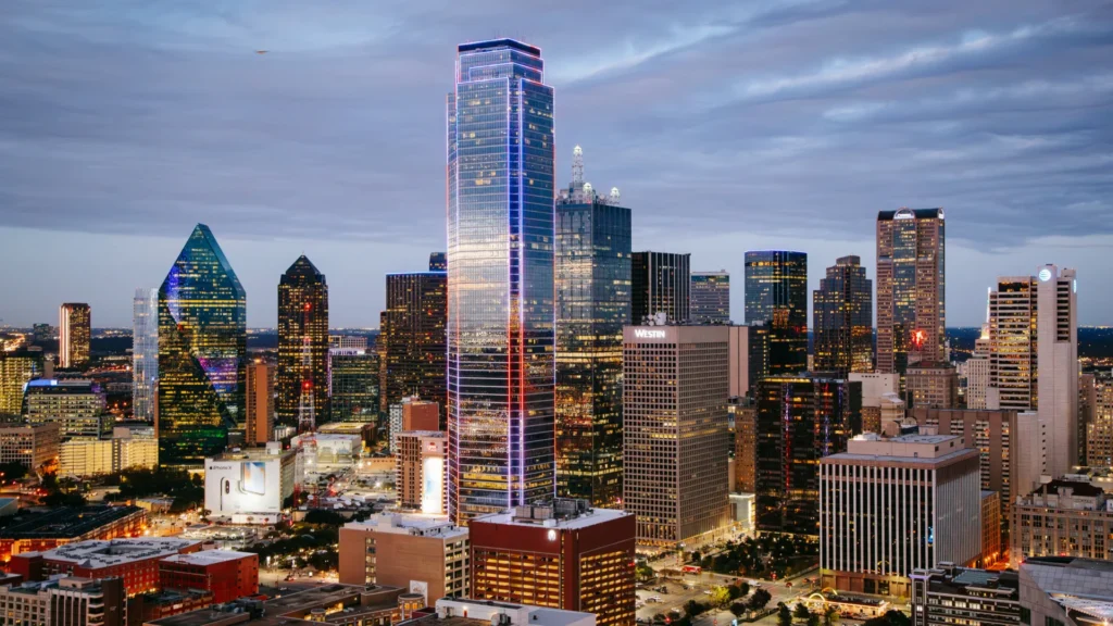 Downtown Dallas skyline at dusk, featuring illuminated modern skyscrapers and high-rise buildings under a cloudy sky.
