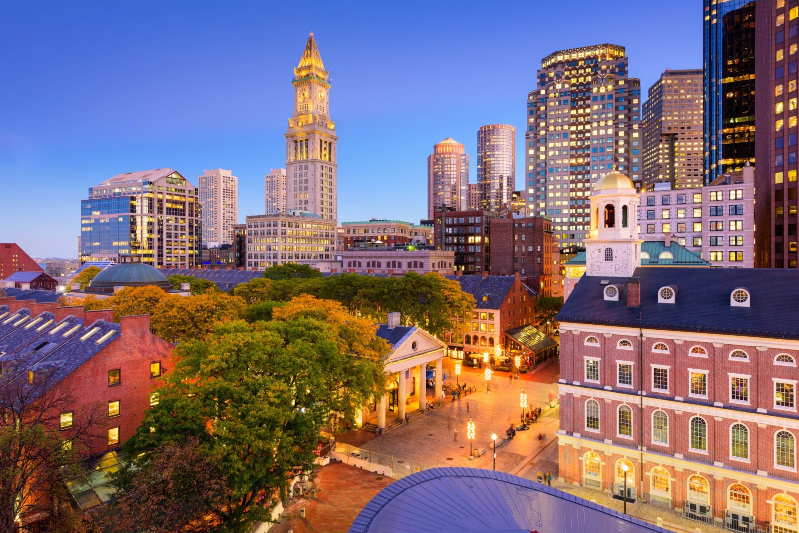 Boston cityscape at dusk featuring Faneuil Hall, Quincy Market, and surrounding modern skyscrapers with trees and illuminated buildings.